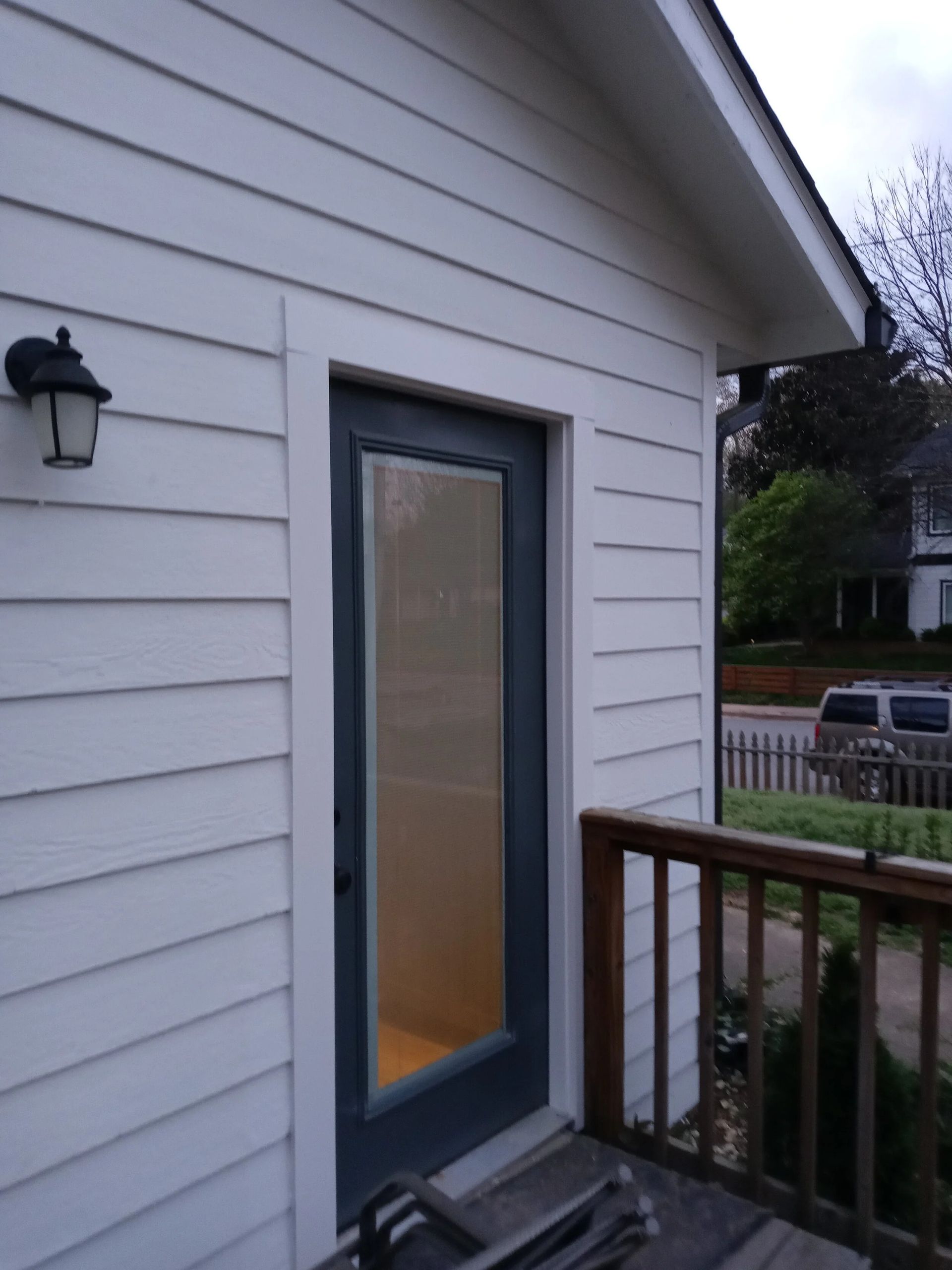 White-sided building with dark-framed door and porch. Door has a long window. Exterior lamp on the side.