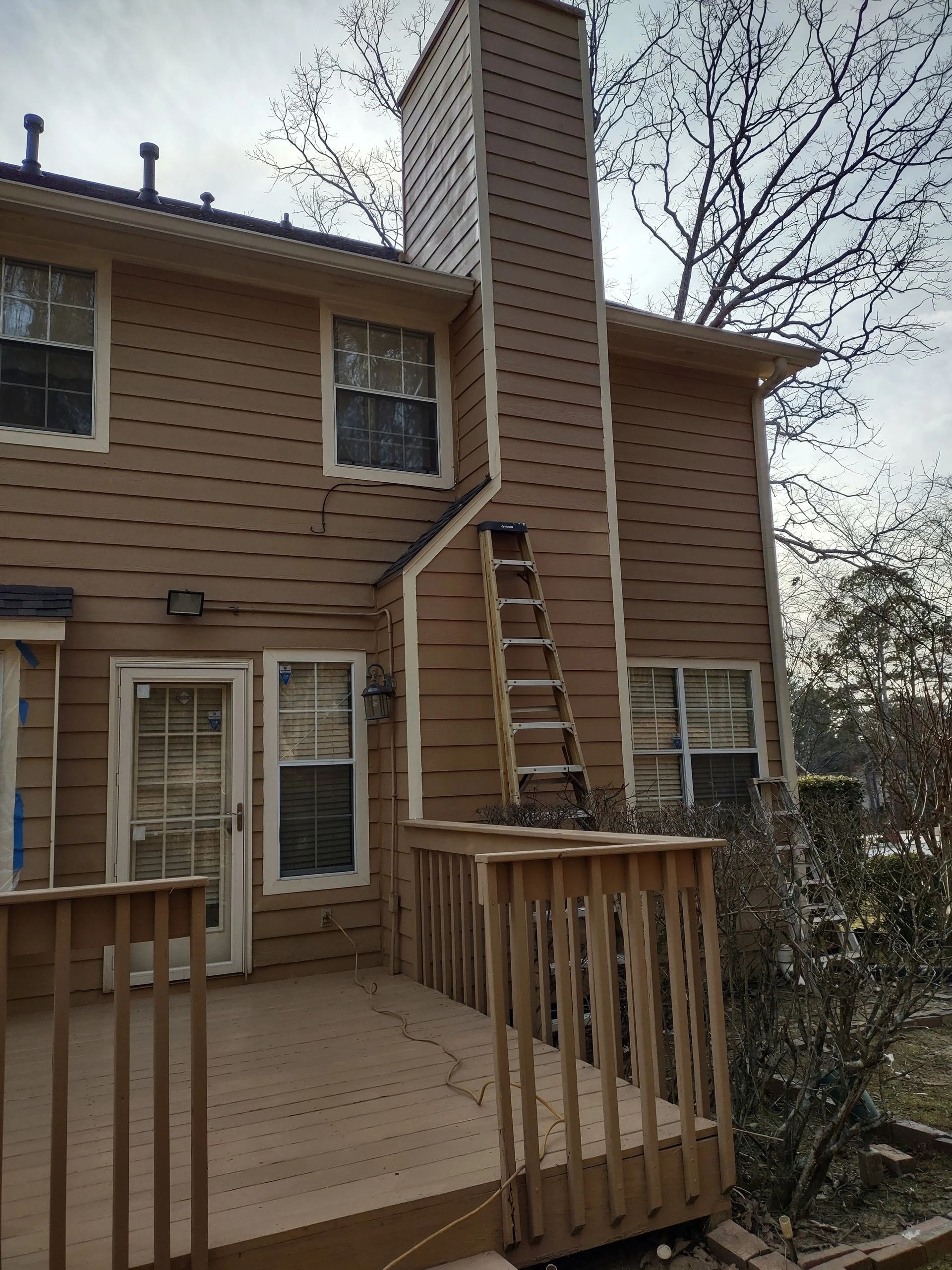House exterior with deck and chimney; ladder leans against chimney. Brown siding, white trim.