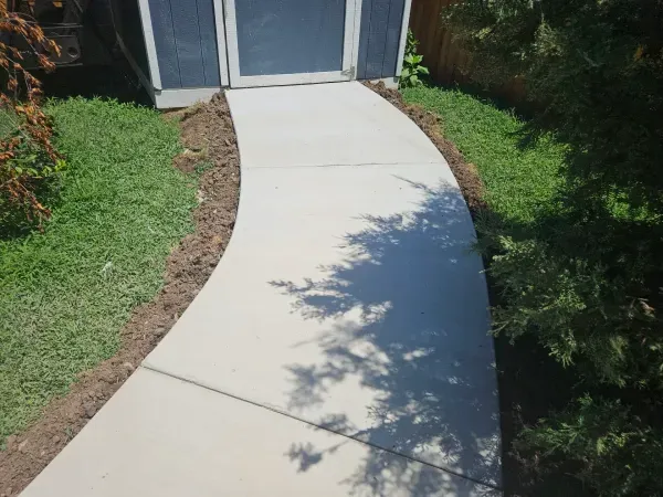 Curved concrete walkway leading to a building entrance, bordered by grass and shrubs. Sunny day.