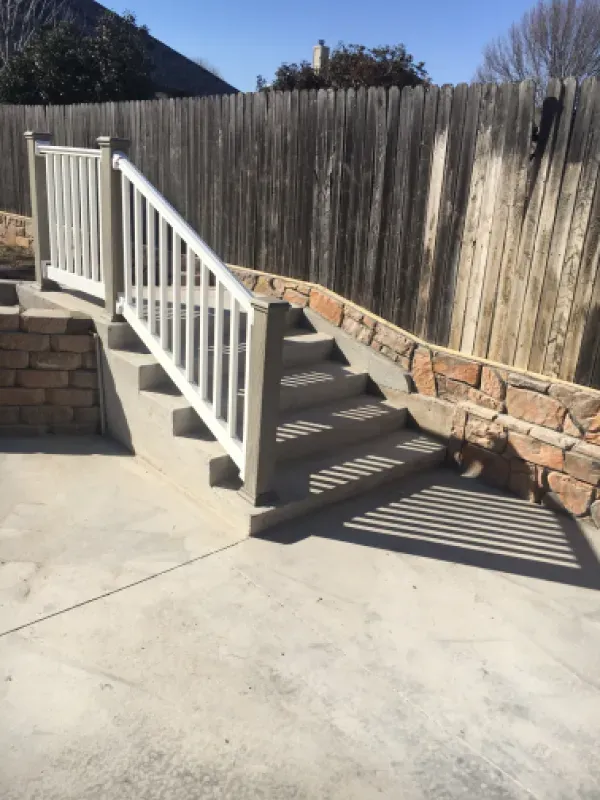 Concrete staircase with white railing, stone wall, and wooden fence. Sunny outdoor setting.