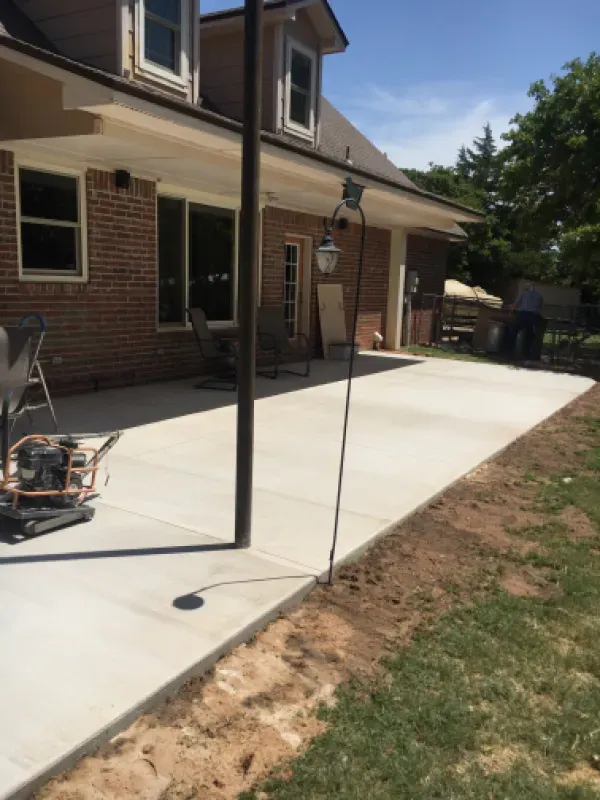 Newly poured concrete patio next to a brick house. A metal pole and construction equipment are visible.