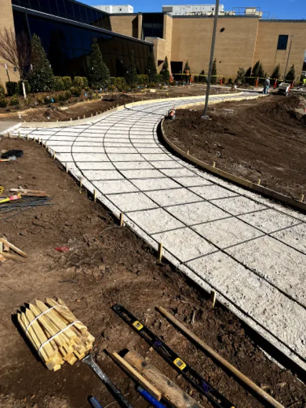 Construction of a curved concrete pathway with rebar grid, surrounded by dirt, near a building.