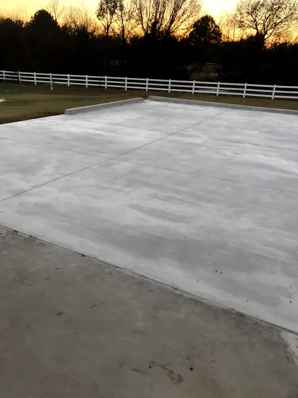 A newly poured concrete slab at dusk, with a white fence and trees in the background.