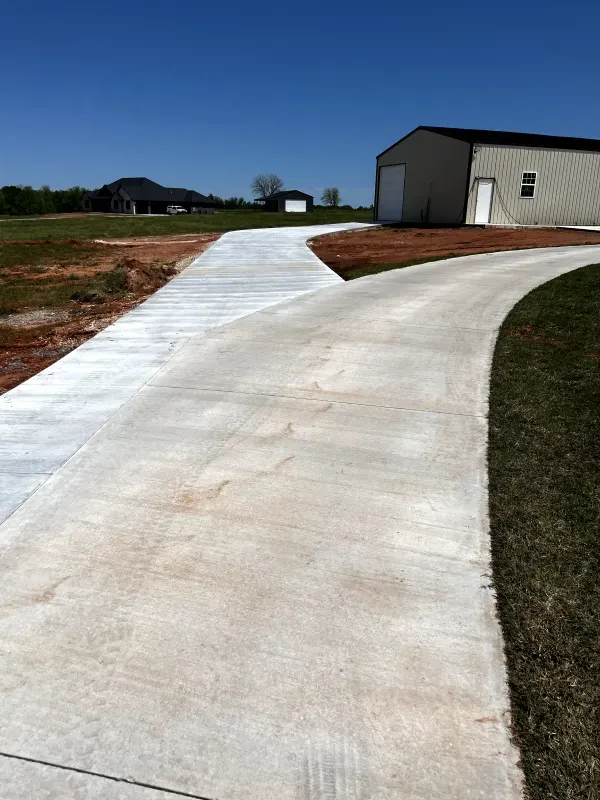 Concrete driveway curves past a metal building and towards a house on a sunny day.