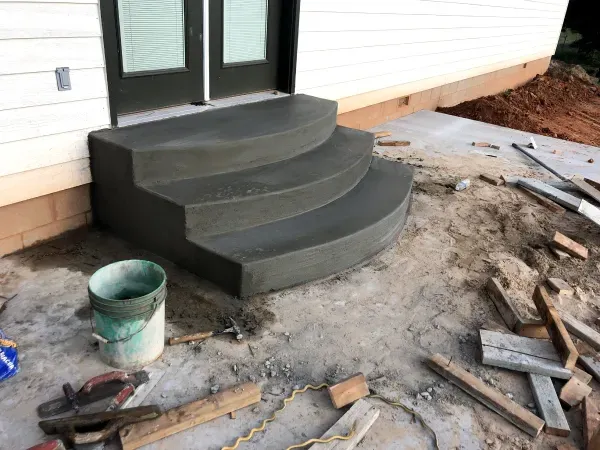 Gray concrete steps leading up to a door, next to a white house, construction materials on the ground.