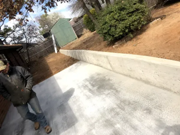 Man stands on a concrete patio next to a concrete retaining wall, a green shed, and a sloped yard.