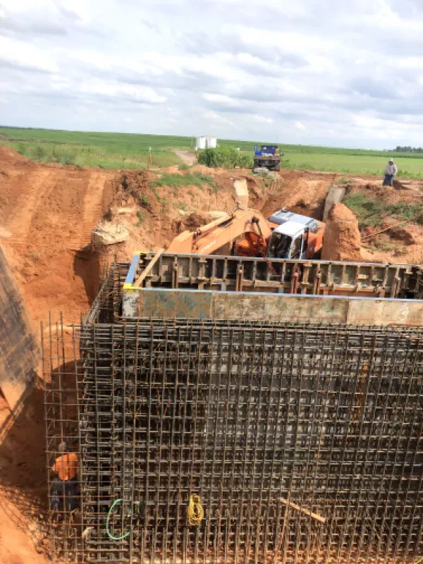 Construction site with rebar framework, earthworks, and machinery. Two workers on site under cloudy skies.