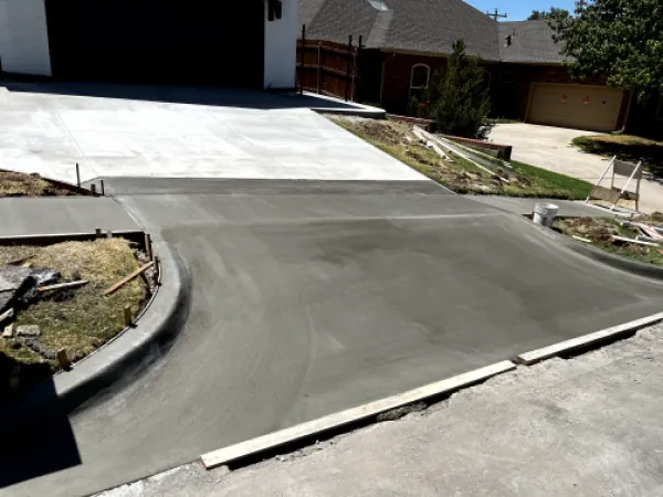 Freshly poured concrete driveway. Gray surface, curved edges, grass borders, house in the background.