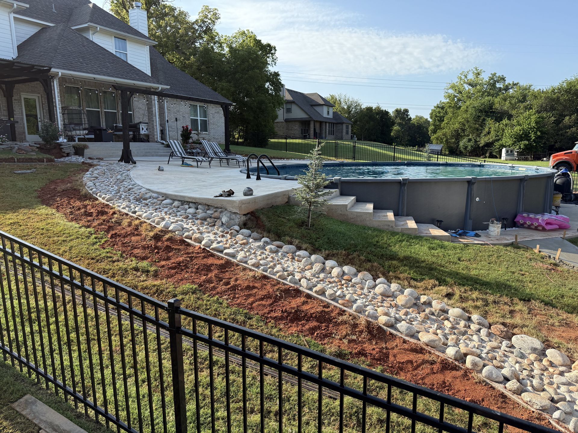 Backyard with a pool, landscaping, house, and black fence under a blue sky.