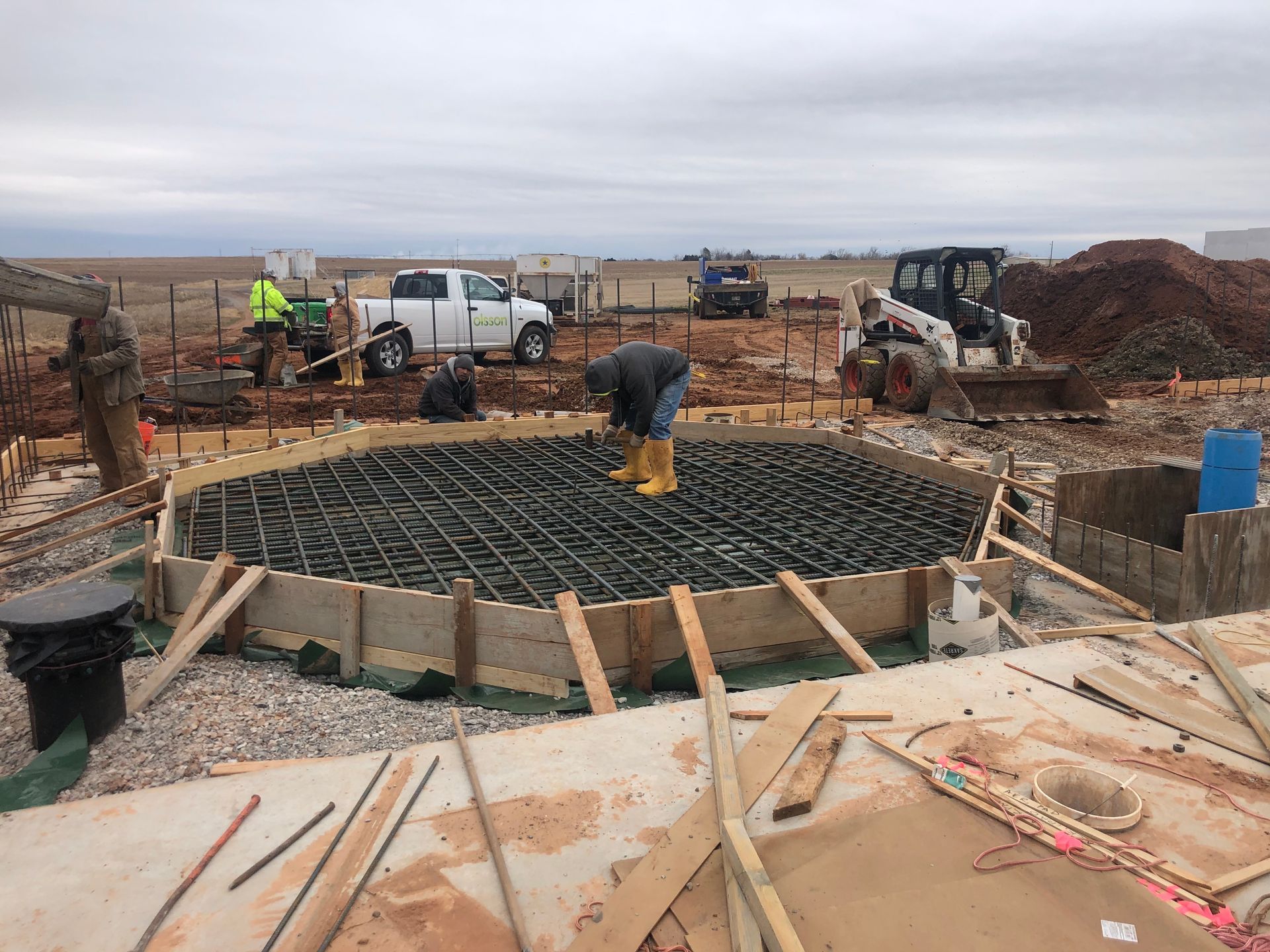 Construction site with workers placing rebar inside a circular concrete form, with trucks and equipment visible.