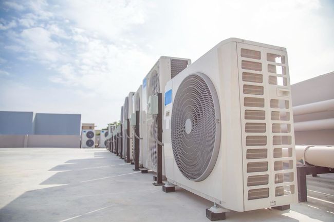 A row of white outdoor HVAC condenser units installed on a flat rooftop against a bright, sunny sky.