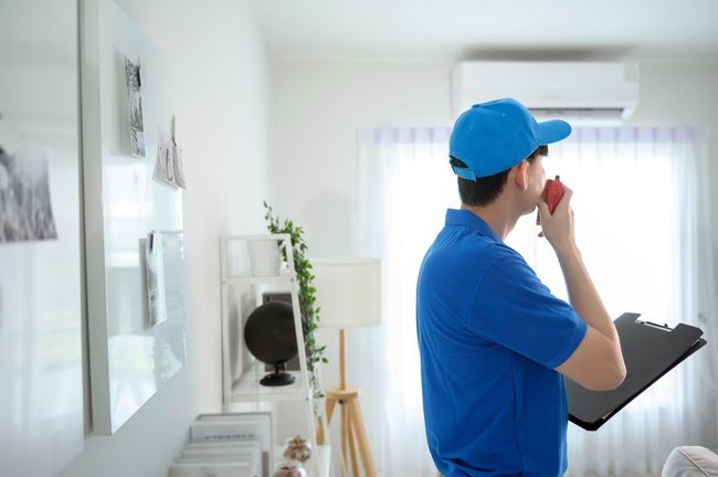 A professional in a blue uniform and cap speaks into a walkie-talkie while holding a clipboard in a bright room.