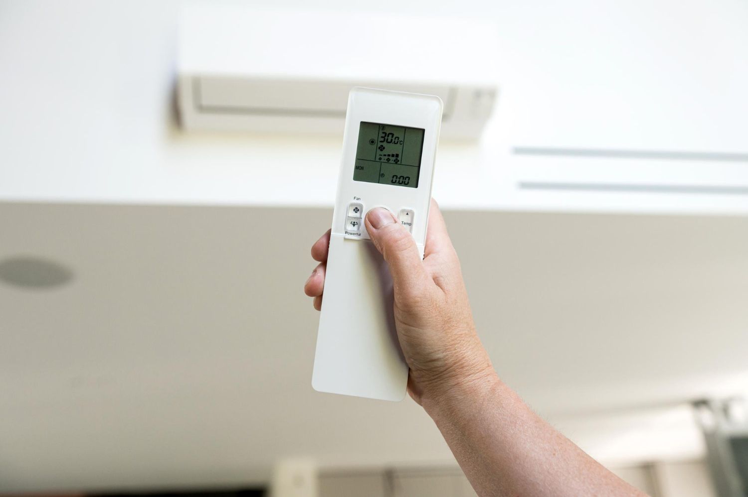 A hand holds a white remote control toward a wall-mounted air conditioning unit in an indoor setting.