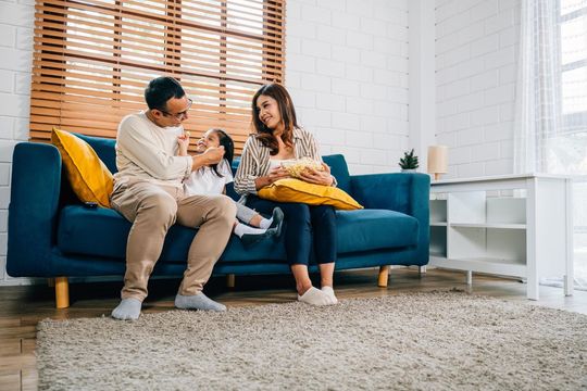 A family sits together on a blue sofa in a brightly lit living room, smiling and interacting with one another.
