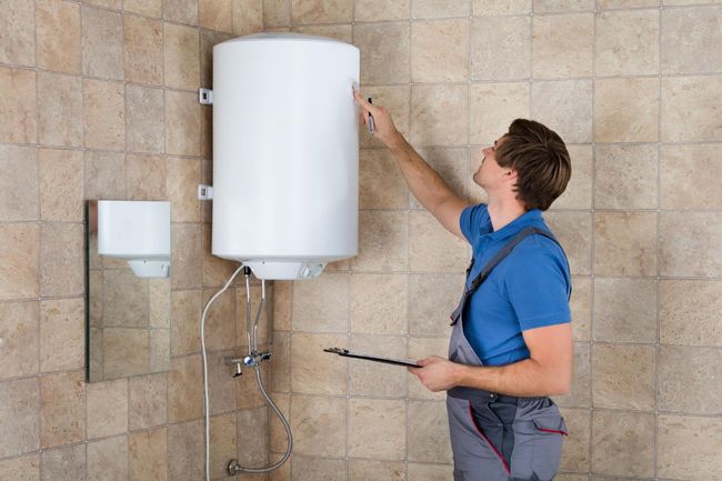 A technician in blue coveralls examines a wall-mounted white water heater in a tiled room, holding a tool in their hand.