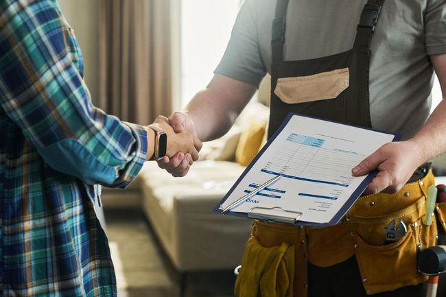 Two people shake hands while one holds a clipboard with a document in a home setting.