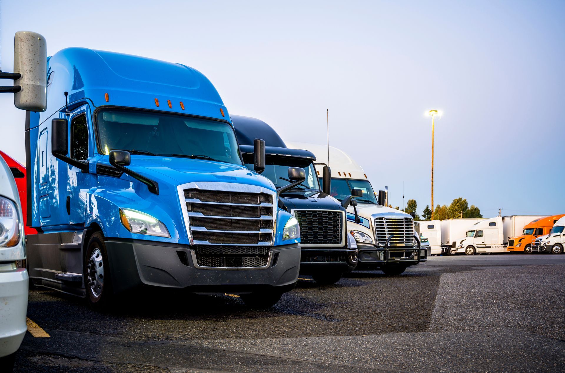 A blue semi-truck is parked in a lot alongside several other trucks under a bright pole light.