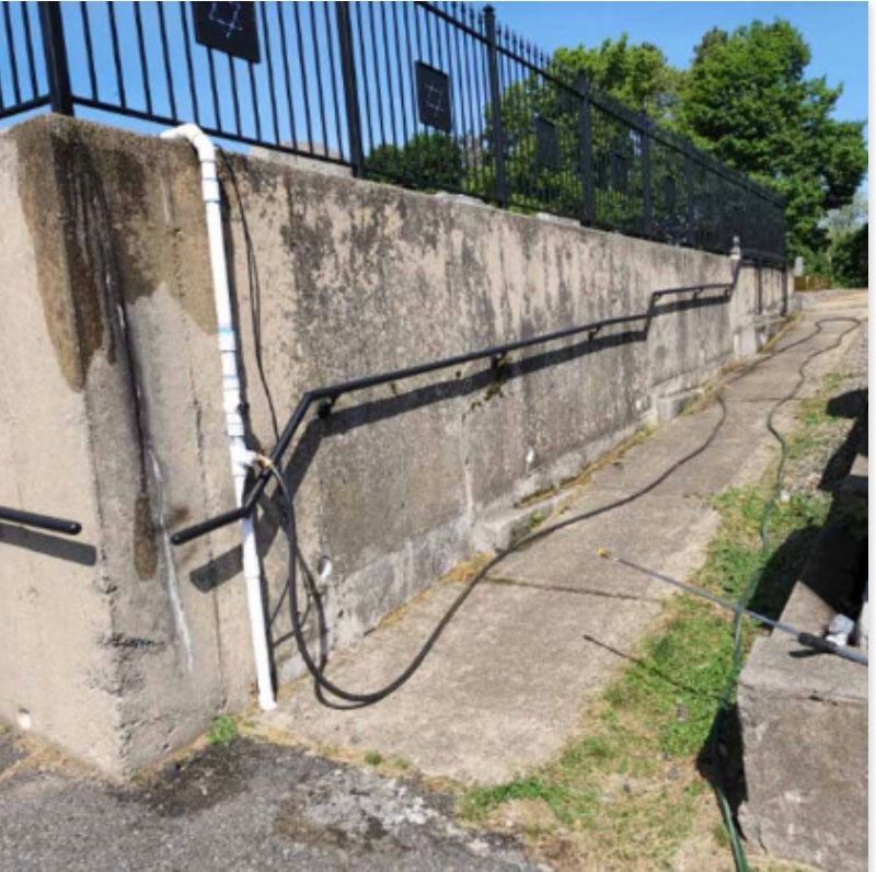 Concrete wall with black railing, black fence in the background, white pipes, and a walkway along the wall.