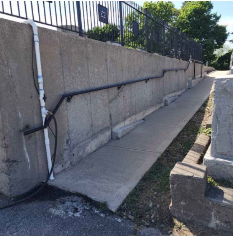 Concrete ramp with handrail alongside a concrete wall, fence overhead.