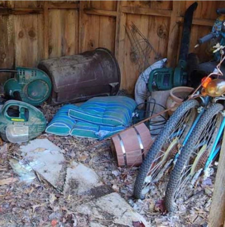 Cluttered shed interior: tools, bike, cushions, containers, and debris on wooden floor.