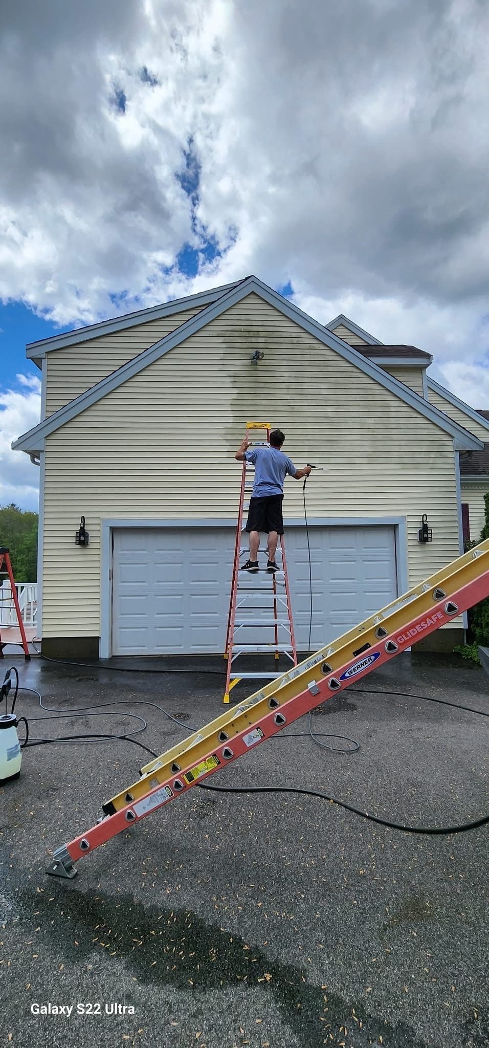A person power washing a house; the left side is cleaned, the right side is dirty. The sky is cloudy.