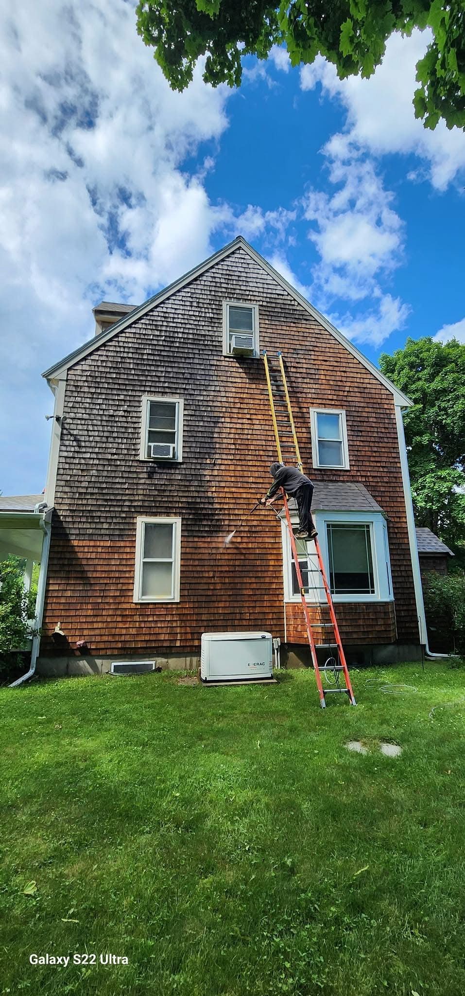 A wooden-shingled house with a ladder, a person on it, and green grass. Blue sky and clouds overhead.