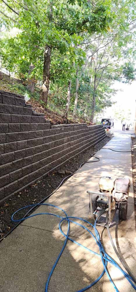 Retaining wall of dark blocks alongside a concrete path with machinery and trees.