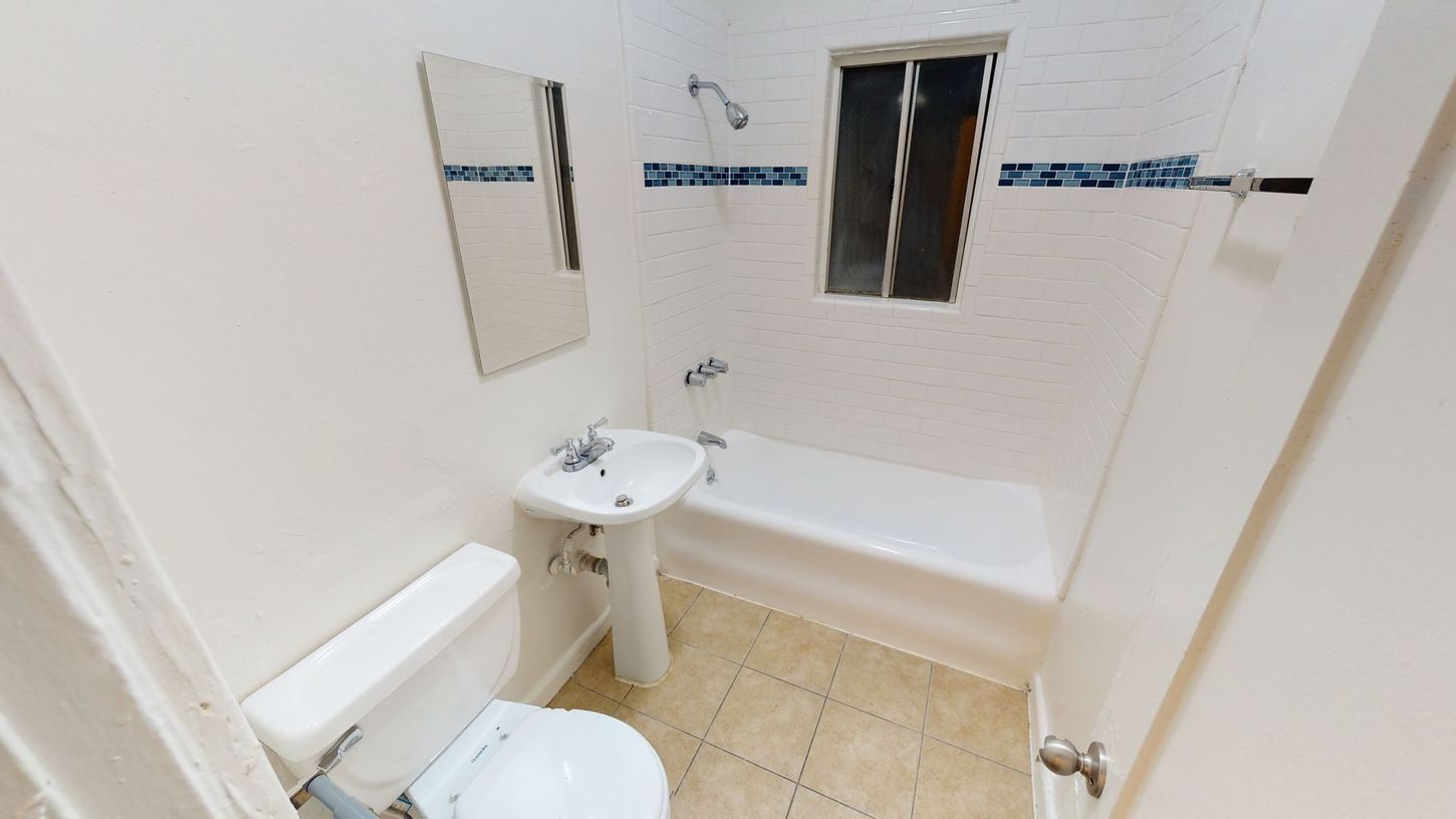 Small bathroom with white walls, toilet, pedestal sink, bathtub, and window. Blue and white tile accents.