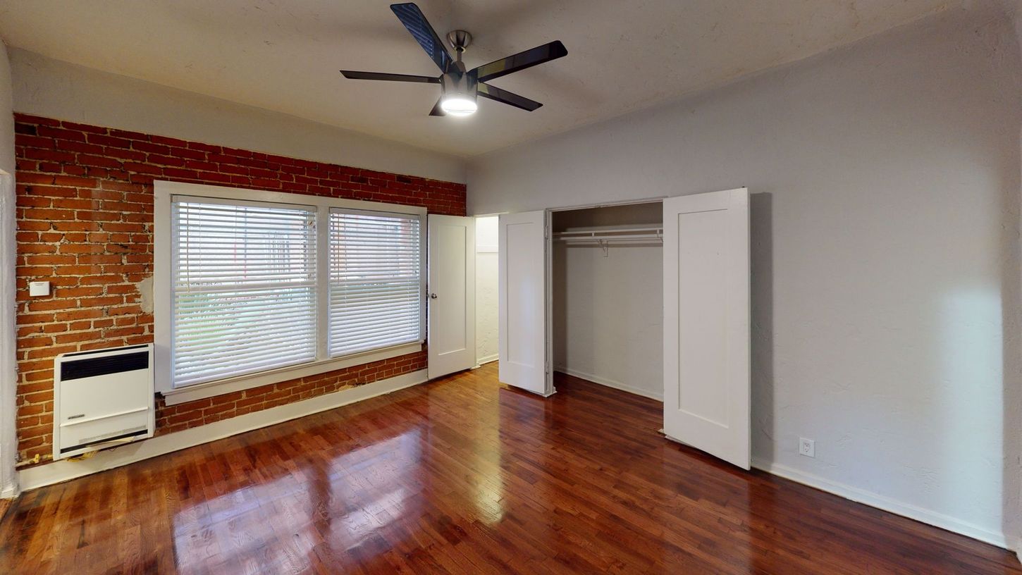 Empty room with hardwood floors, exposed brick wall, window, closet, and ceiling fan.