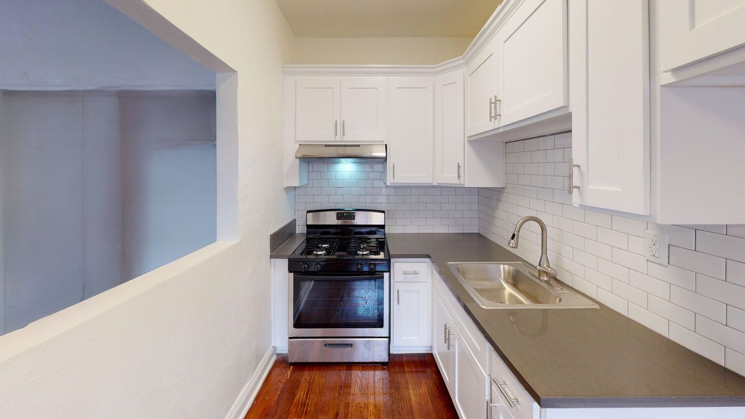 Small, white kitchen with gray countertops, stainless steel appliances, and wood flooring.