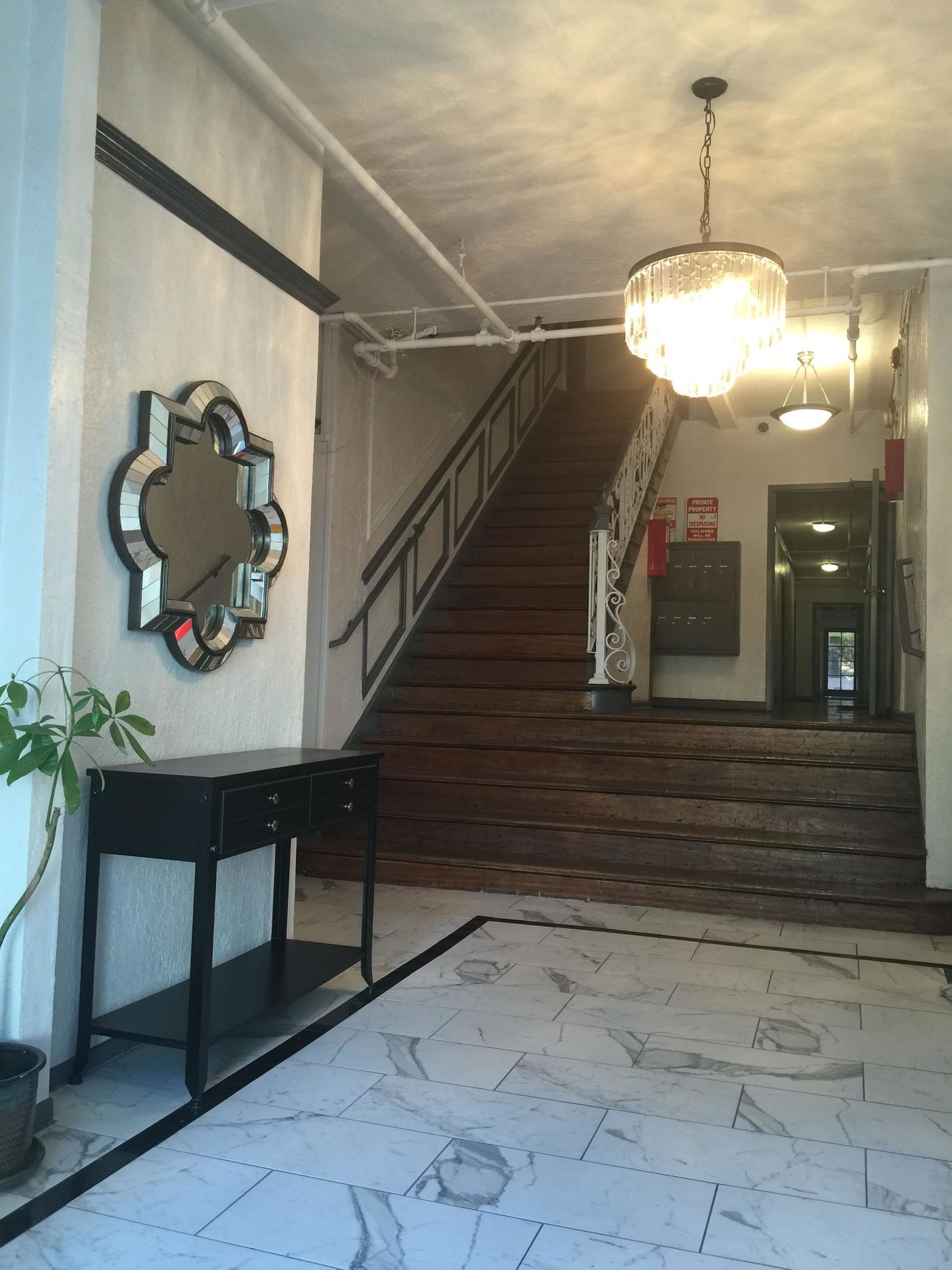 Hallway with staircase, ornate mirror, black table, and chandelier. Marble floor, white walls.