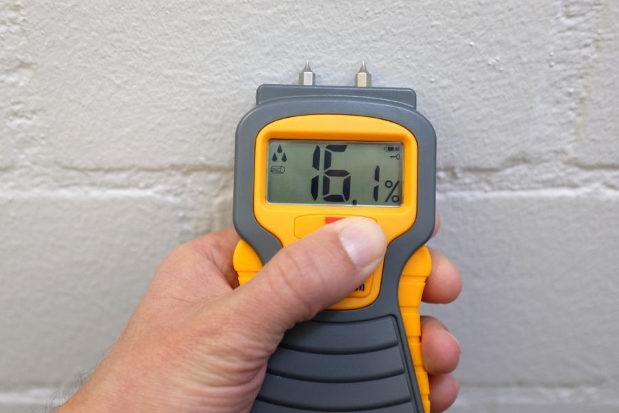 Moisture meter displaying 16.1% pressed against a painted block wall during water damage assessment