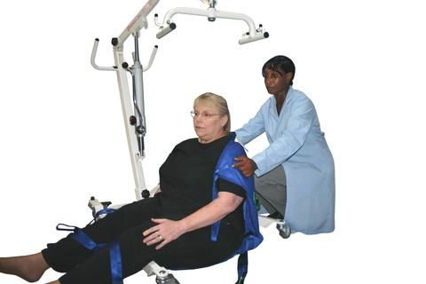 Woman in sling being assisted by caregiver in a medical setting, lift equipment visible.