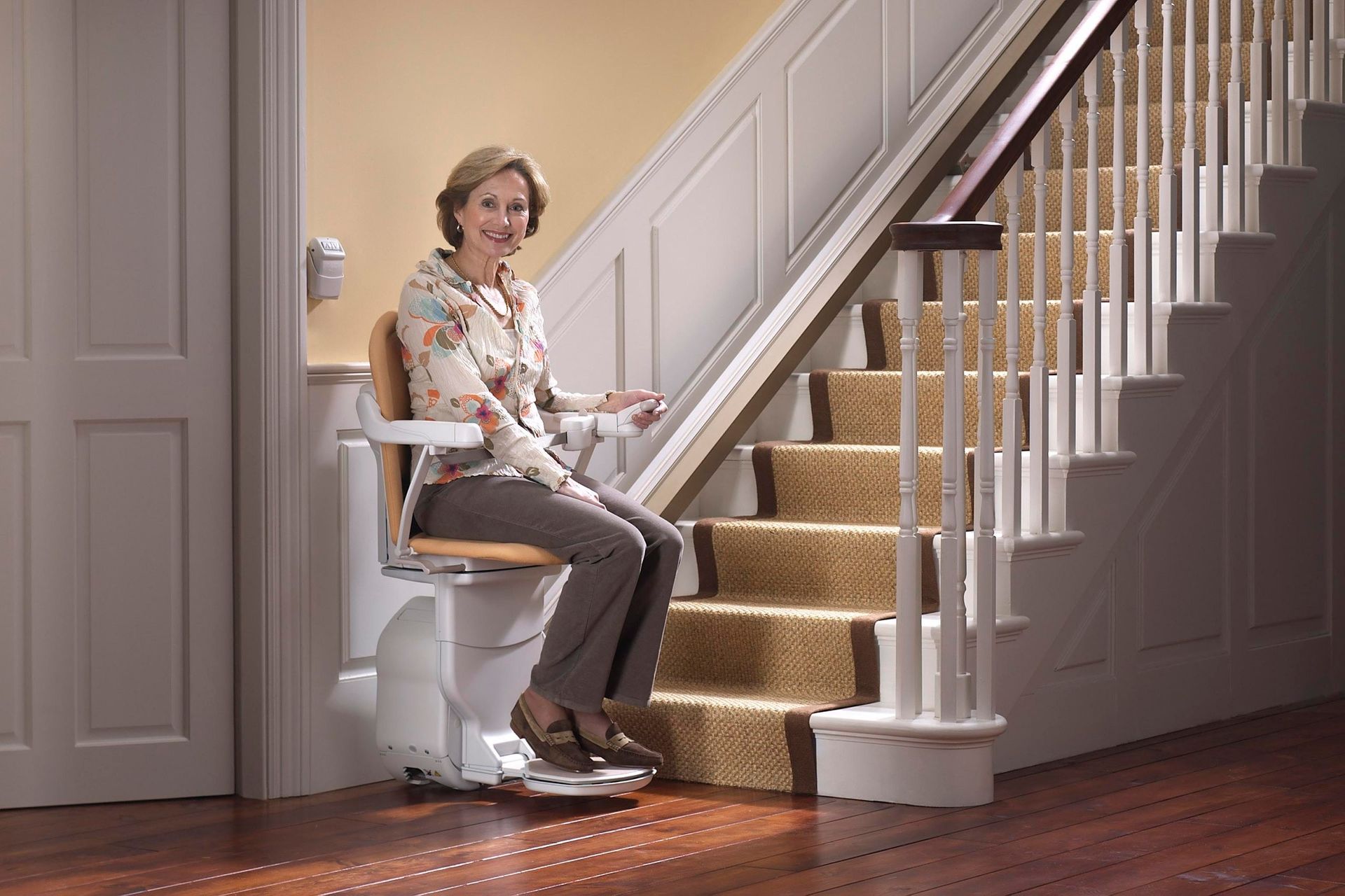 Woman on a stairlift smiles while ascending stairs in a home. Beige carpet and light wooden trim are visible.