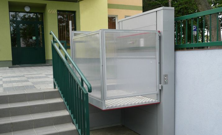Wheelchair lift next to building entrance with stairs, green railings, and a metal platform.