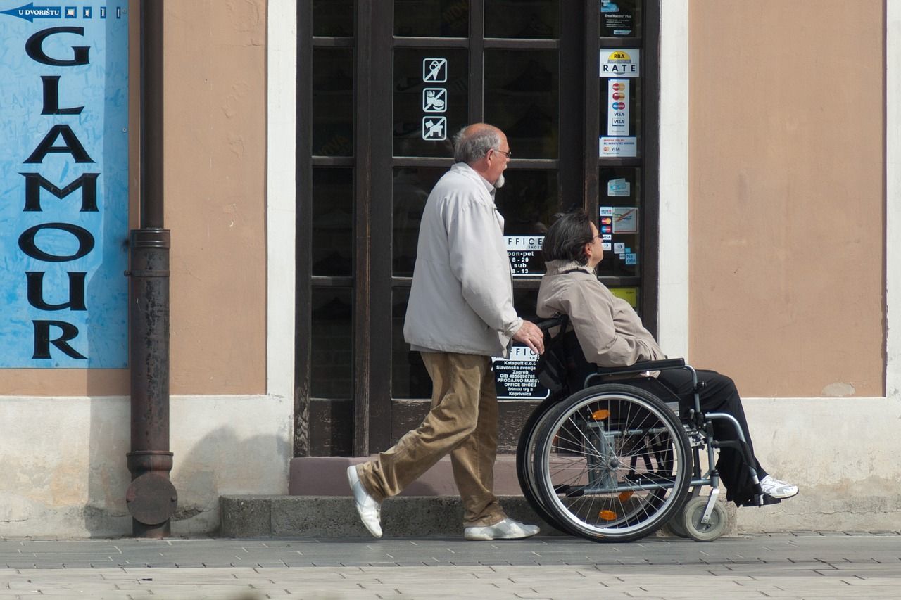 Man pushing a person in a wheelchair on a sidewalk, near a building with a 