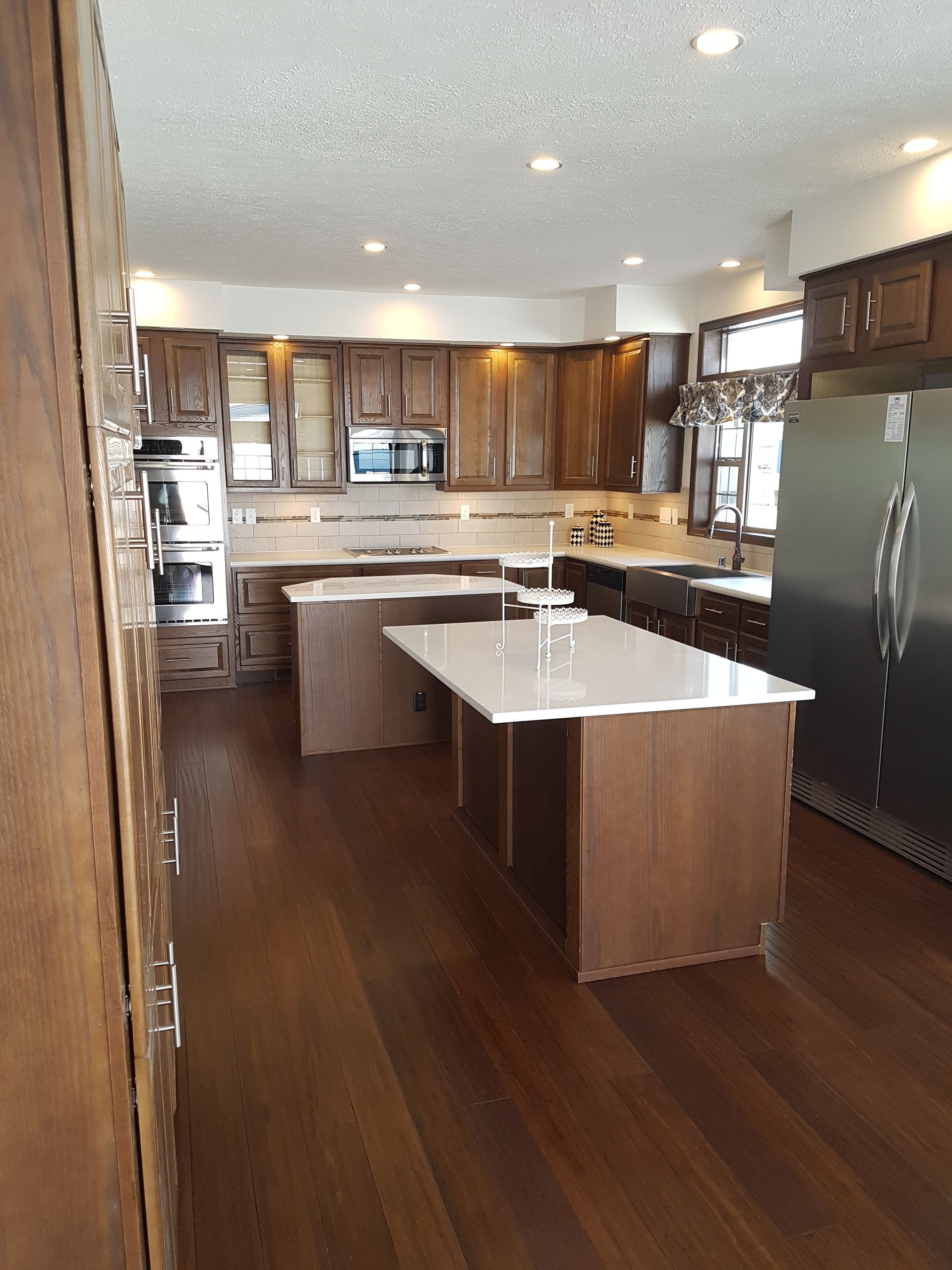 Spacious kitchen with wooden cabinets, two islands with white countertops, stainless steel refrigerator.