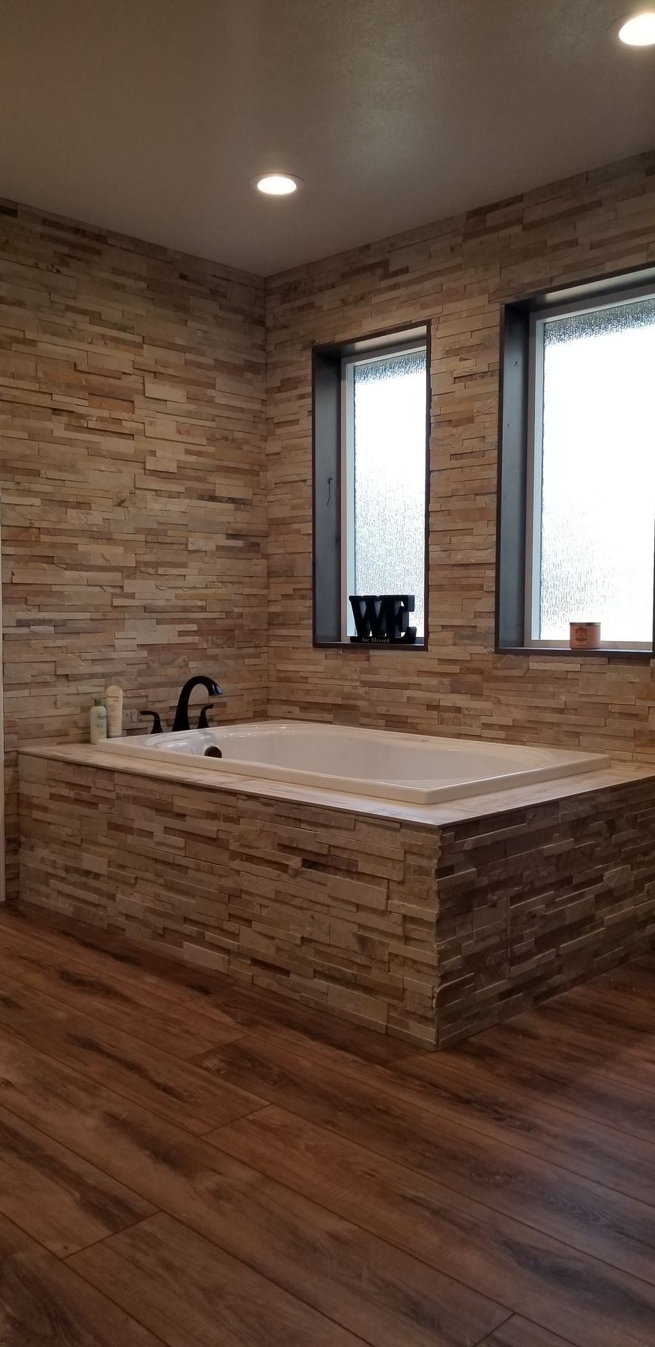 Bathroom with stone-covered walls and a built-in tub, with wooden floor. Sunlight streams through the windows.