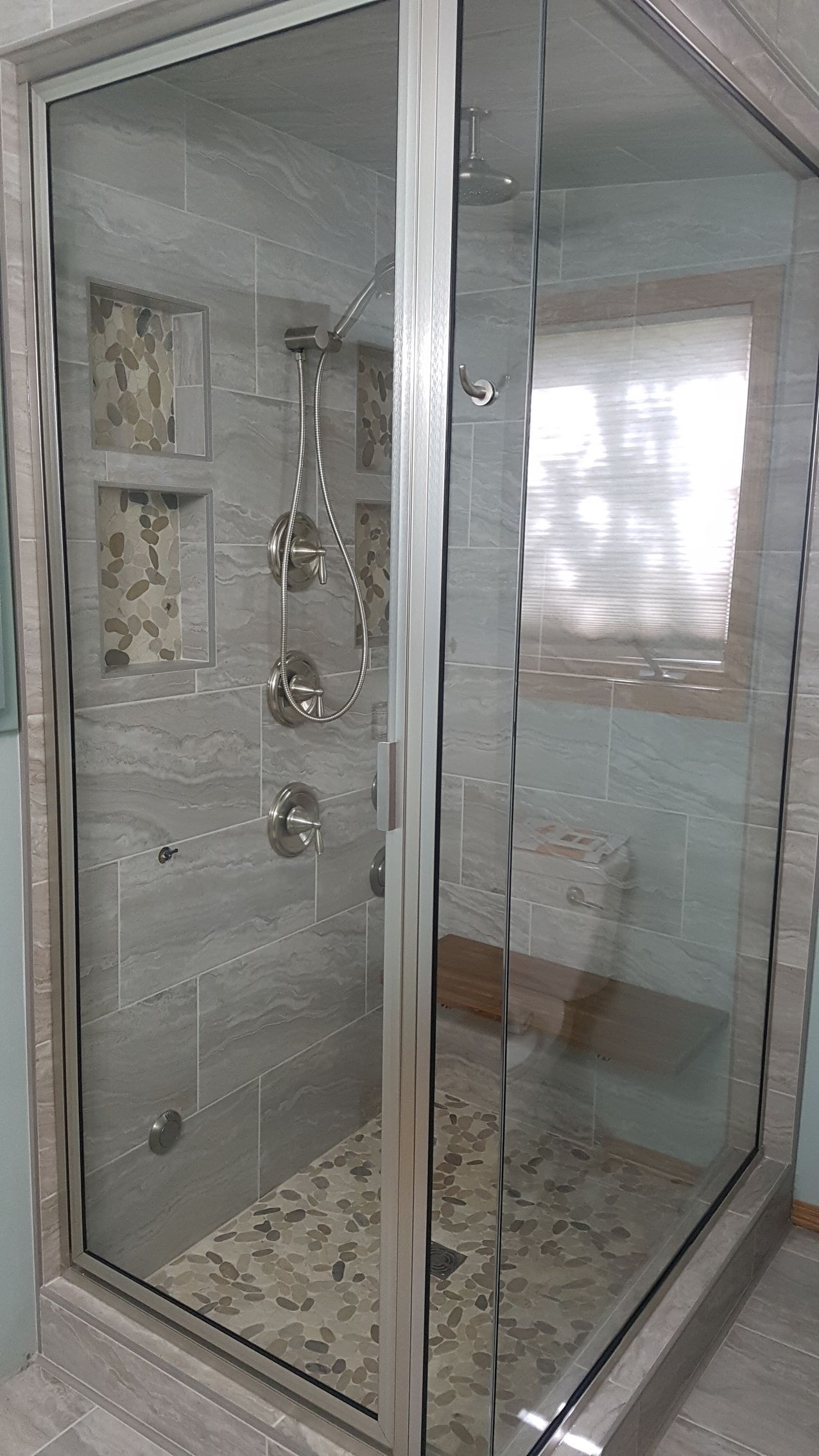 Glass-enclosed shower with gray tile walls and a pebble floor. Chrome fixtures and a small window are visible.