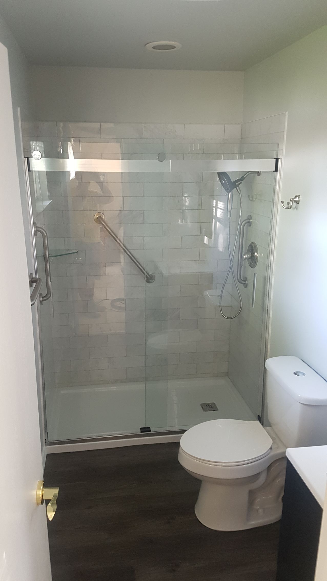 Bathroom with glass shower, toilet, and dark flooring. Handrail visible in the shower.
