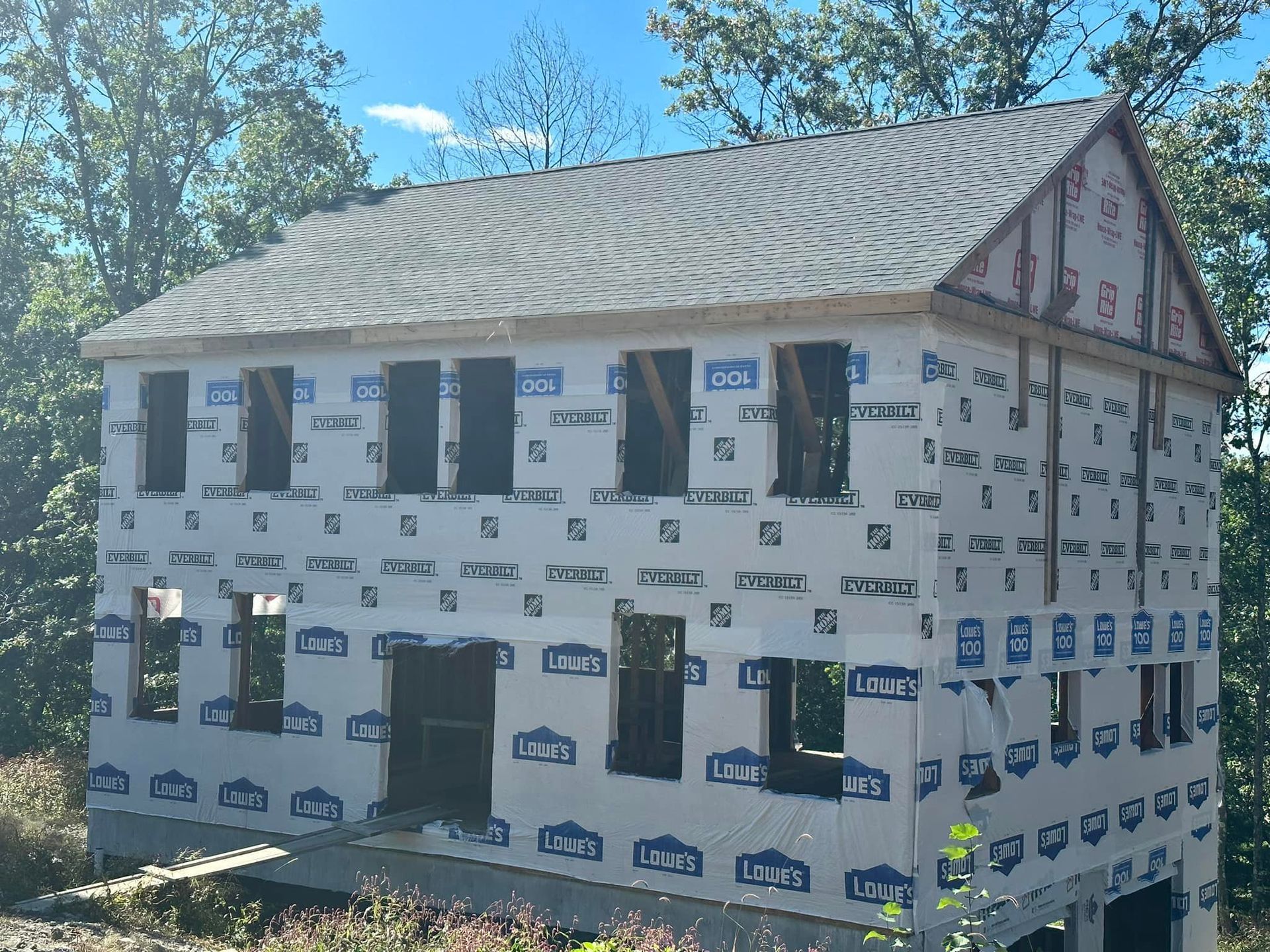 Two-story house under construction, sheathing and dark roof, set in a wooded area.