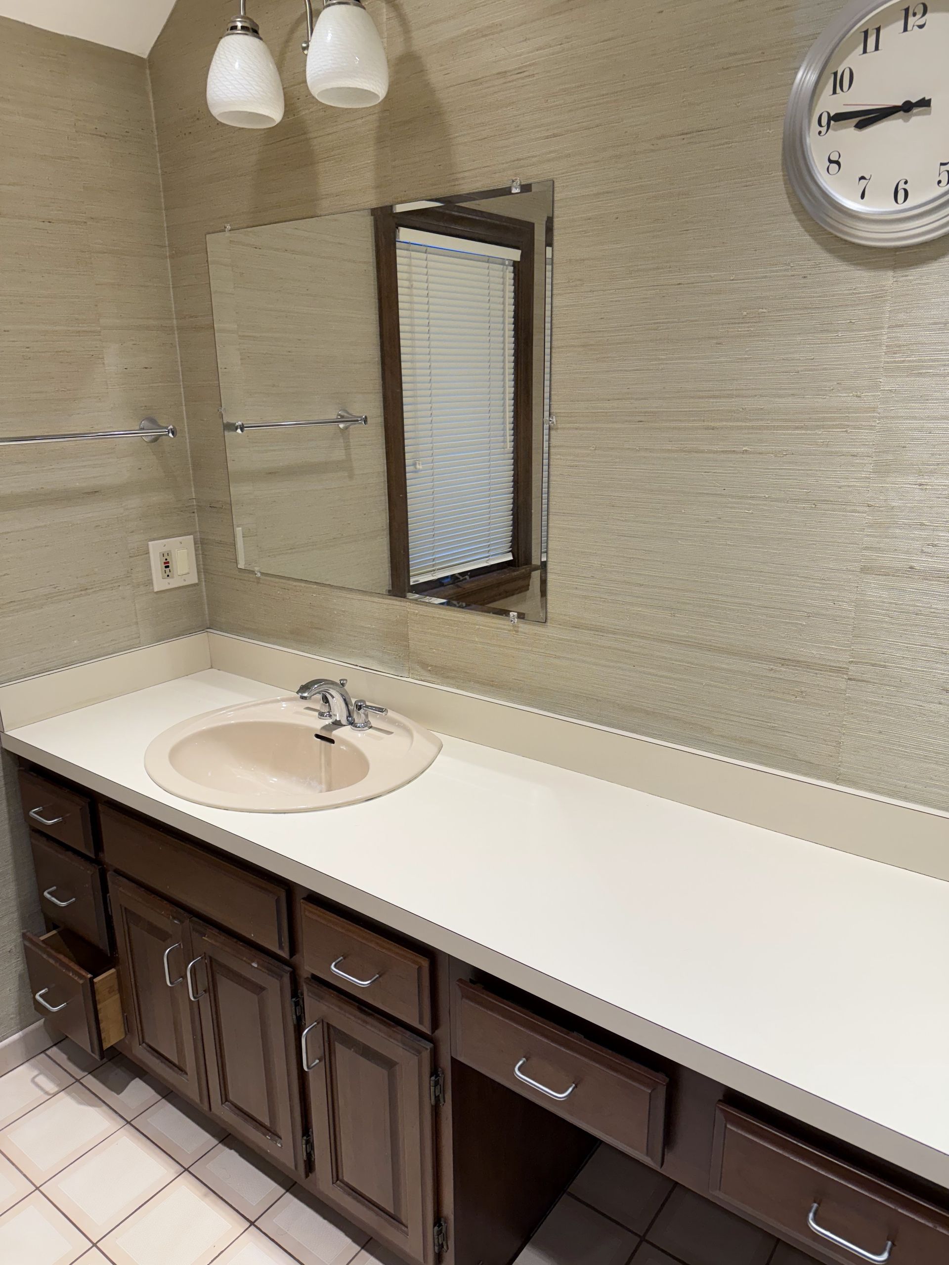 Bathroom with a vanity, mirror, clock, and two light fixtures. Beige walls, white countertop, dark cabinets.