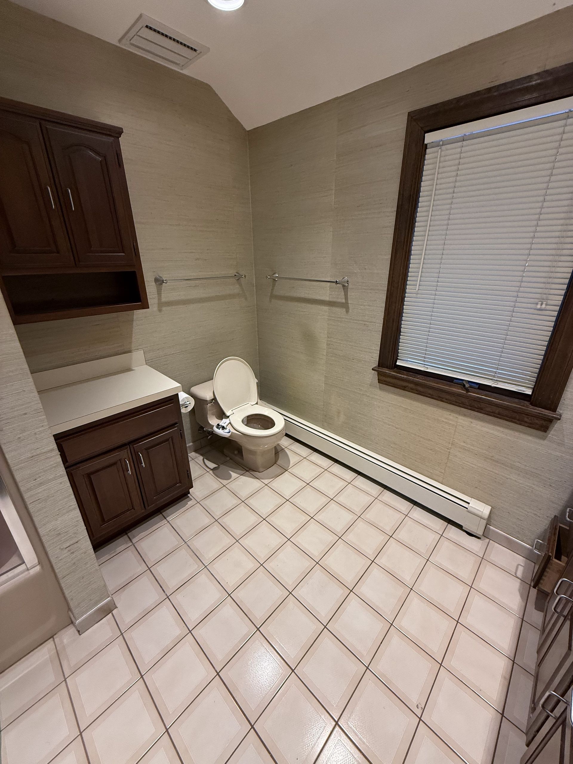 Bathroom with brown cabinets, toilet, and window with blinds. Tile floor and neutral-toned walls.