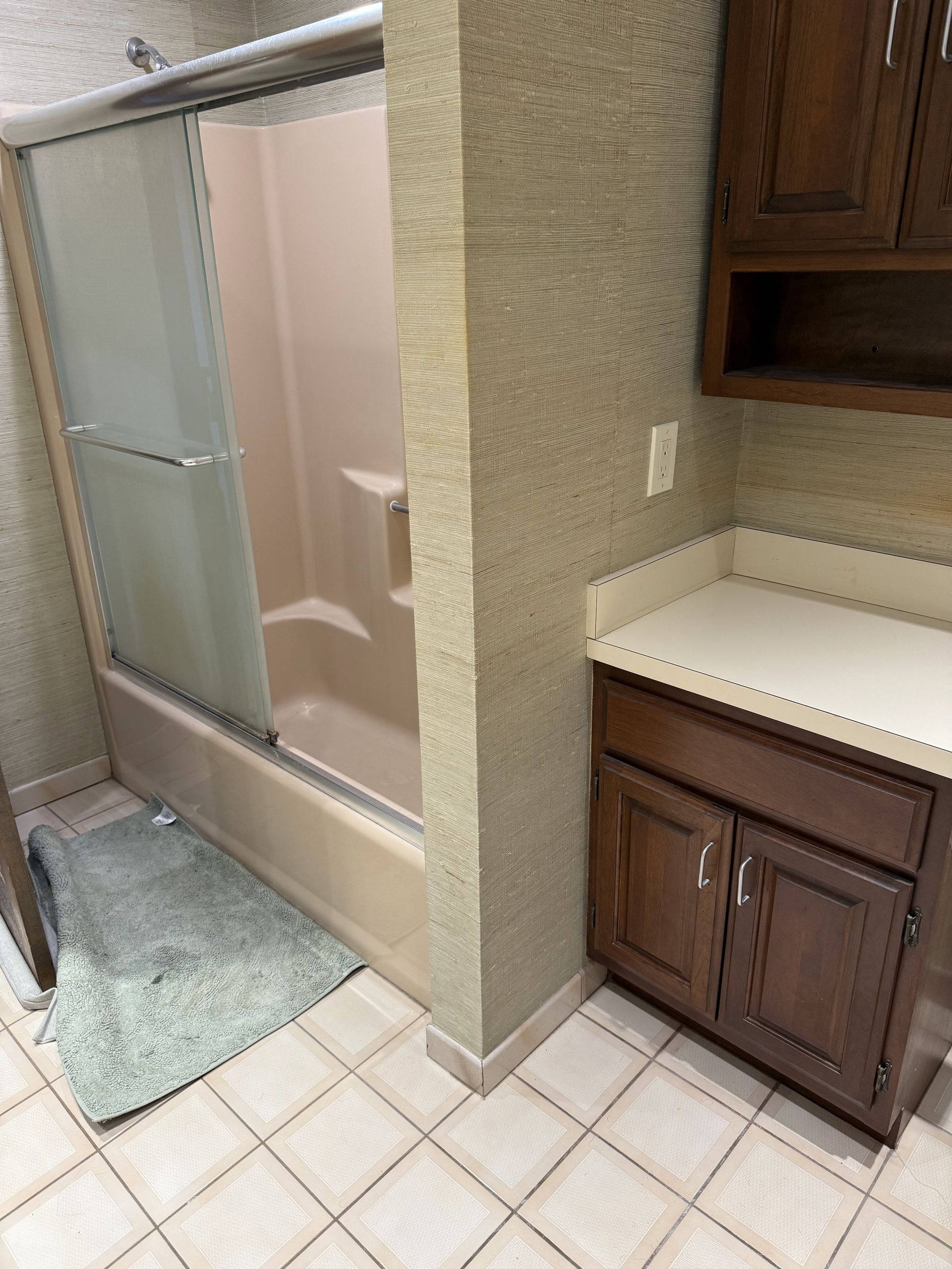 Bathroom with shower, vanity, and brown cabinets; tan shower, light-colored tile flooring, green bath mat.