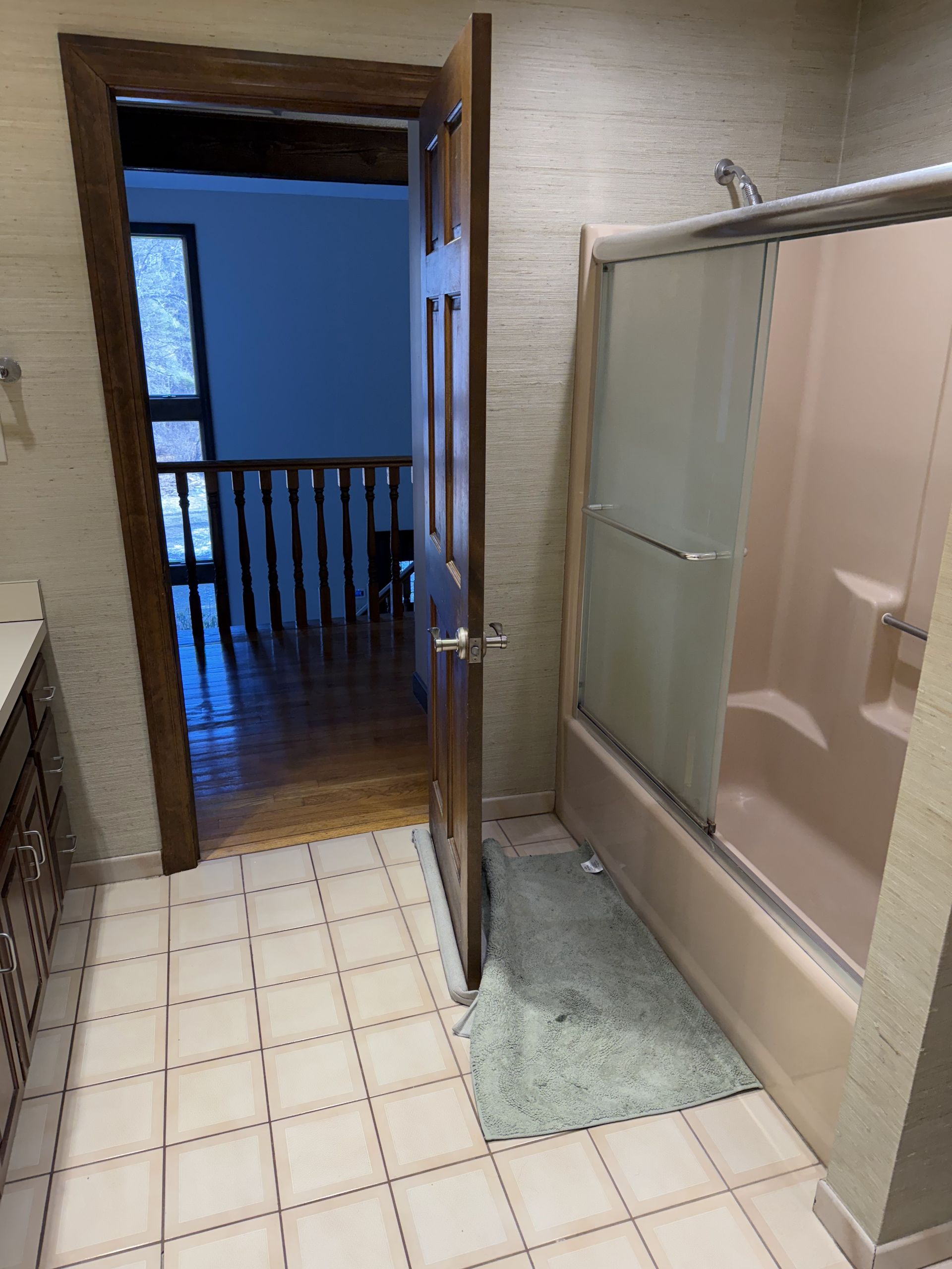 Bathroom with open wooden door, shower, and tile floor, leading to a hallway with stairs.