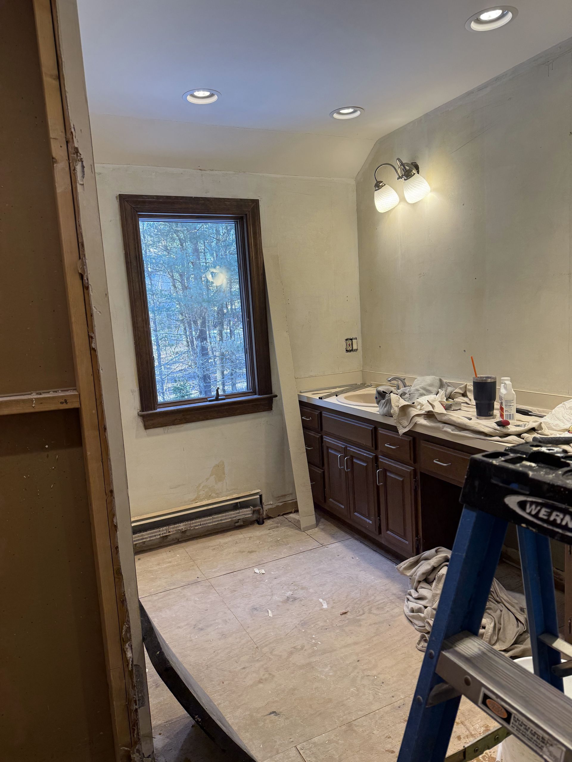 Bathroom under renovation, featuring a window, vanity, and a ladder. Beige walls, brown cabinets.