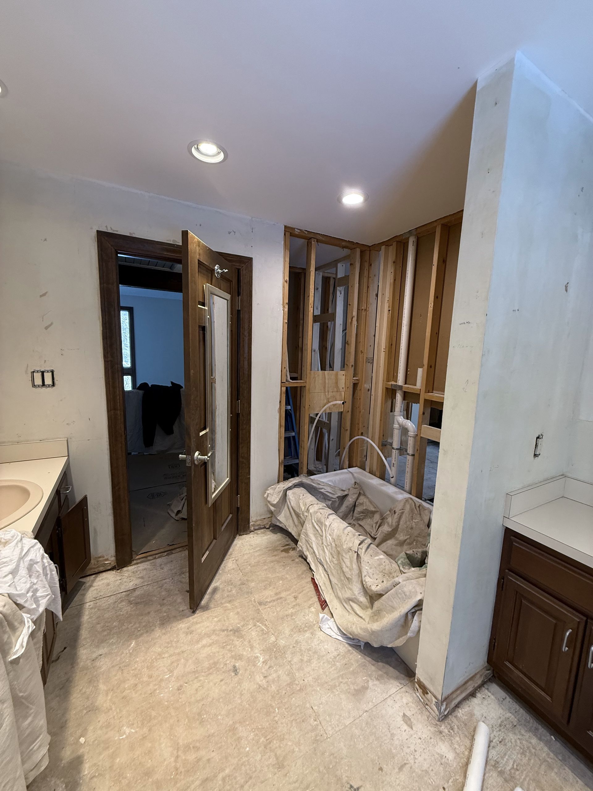 Bathroom renovation, unfinished walls, partially covered bathtub, doorway, cabinets on either side.