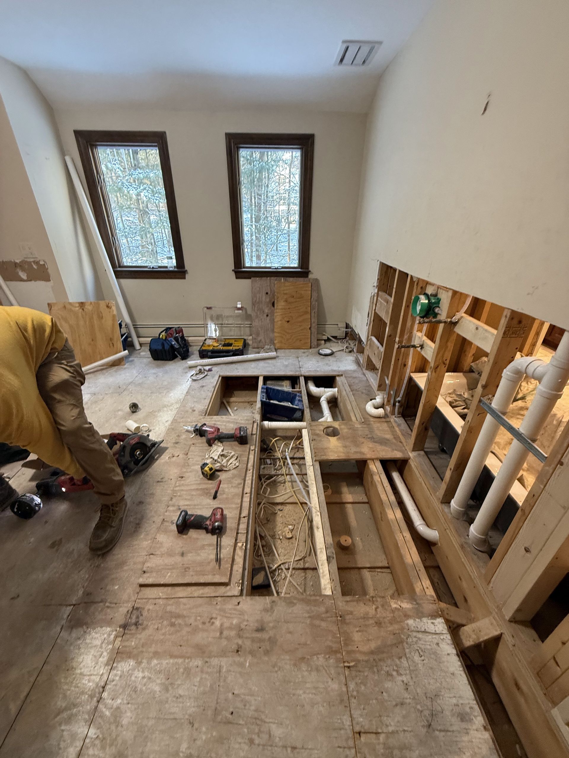 Room renovation with exposed plumbing and flooring. Person working, walls open. Windows visible.