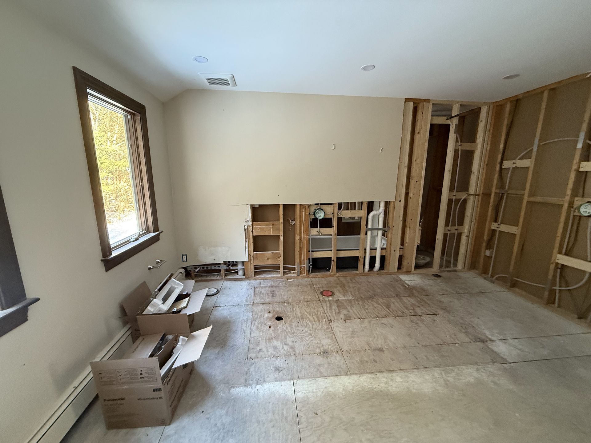 Interior room under renovation, exposed framing, drywall removed. Boxes, window, and flooring visible.