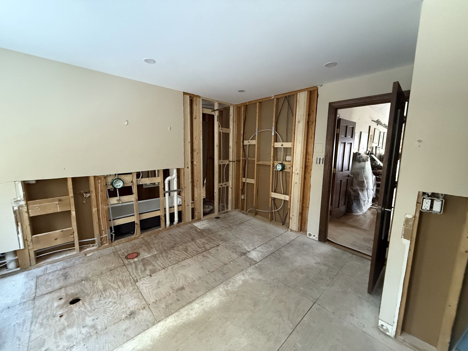 Room undergoing renovation, walls open to studs, plumbing and wiring exposed. Doorway and drywall debris visible.